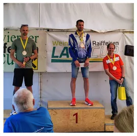 Drei Männer stehen auf einem Podium mit Medaillen um den Hals. Ein Mann hält ein Papier und trägt Turnschuhe. Die anderen zwei tragen Freizeitkleidung. Hinter ihnen ist ein Banner mit dem Text Wein LA und Raiffeisen Cup.