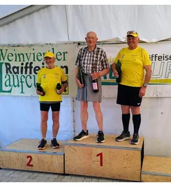 Drei ältere Männer stehen auf einem Podium, halten Flaschen in der Hand, wahrscheinlich bei einer Weinprobe. Der Mann links trägt ein gelbes Shirt, der mittlere Mann ein kariertes Shirt und der rechte Mann ebenfalls ein gelbes Shirt. Sie stehen auf einer hölzernen Plattform mit den Nummern 1, 2 und 3.