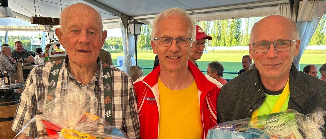 Drei Männer, einer mit Geschenkkorb, einer mit Snackkorb, stehen unter einem Zelt. Einer trägt eine Brille und eine Jacke.
