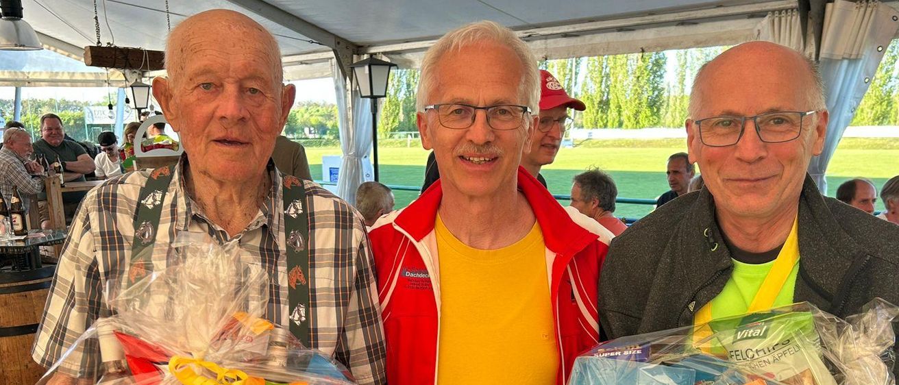 Drei Männer, einer mit Geschenkkorb, einer mit Snackkorb, stehen unter einem Zelt. Einer trägt eine Brille und eine Jacke.