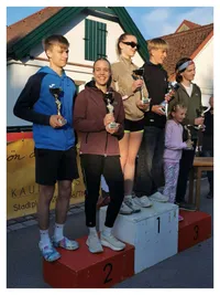Sechs Personen stehen auf einem Podest und halten Trophäen. Eine Frau in der Mitte trägt eine braune Jacke und lächelt. Dahinter halten zwei Frauen Trophäen. Ein gelber Banner ist im Hintergrund.