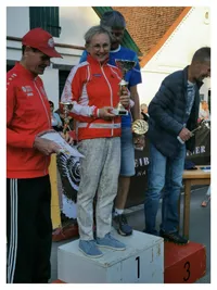 Eine Gruppe von Menschen steht auf einem Podest. Die Frau in der Mitte hält eine Trophäe. Der Mann auf der linken Seite hält ein Stück Papier. Der Mann auf der rechten Seite schaut die Frau an. Hinter ihnen sind ein Tisch und ein Gebäude.