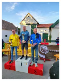 Drei Männer stehen auf einem Podium und halten Trophäen, zwei links in Gelb und Blau, einer rechts in Blau. Ein Banner mit einem Spirallogo ist hinter ihnen.