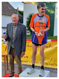 Zwei ältere Männer stehen vor einem Banner, einer in einem grauen Anzug, der einen Pokal hält, der andere in einem blau-orangefarbenen Sporttrikot, das einen Pokal hält.