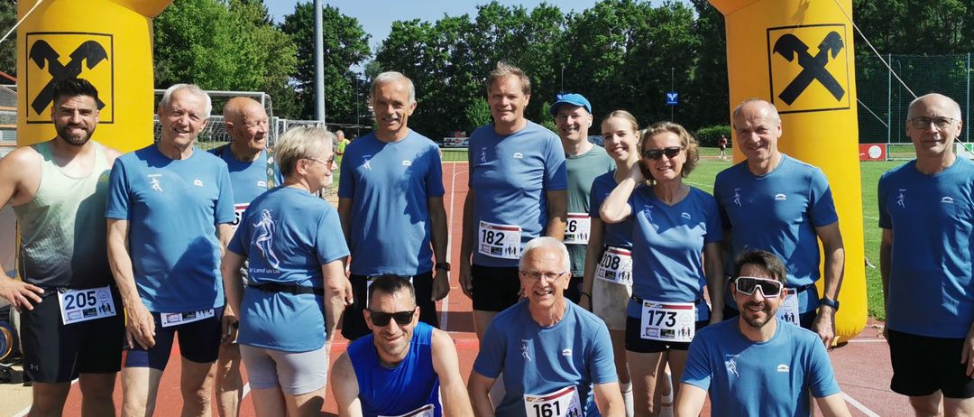 Eine Gruppe von Läufern, die blaue T-Shirts mit Nummern tragen, posiert für ein Foto vor einem gelben Bogen mit Raiffeisen. Bäume und ein blauer Himmel sind im Hintergrund sichtbar.