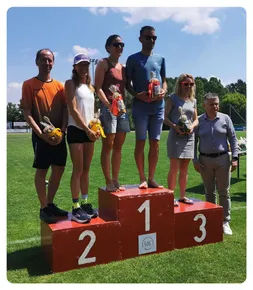 Eine Gruppe von Athleten steht auf einem roten Podest, hält Geschenktüten in den Händen, wobei einer eine Sonnenbrille trägt und die beiden anderen Frauen Turnschuhe tragen.