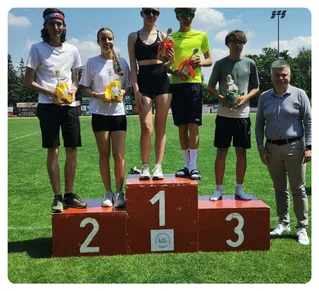 Fünf Personen stehen auf einem Podest mit den Nummern 1, 2 und 3. Sie halten Pokale und tragen lässige Sportkleidung. Der Mann rechts trägt einen Gürtel und ein Poloshirt. Die Frau in der Mitte trägt ein schwarzes Crop-Top und Shorts. Der Hintergrund besteht aus einem Rasenfeld und einem Stadion mit Bäumen.