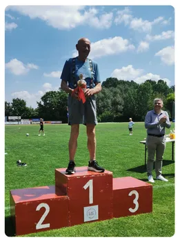 Ein Mann steht auf einem roten Podest und hält einen Pokal. Er lächelt einen Mann an, der applaudiert. Im Hintergrund befindet sich ein Feld mit spielenden Kindern und Bäumen.