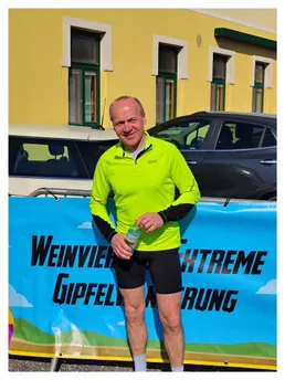 Ein Mann steht vor einem gelben Gebäude und hält eine Wasserflasche in der Hand. Er trägt ein neongrünes Jackett und schwarze Shorts. Hinter ihm steht ein blauer Banner mit der Aufschrift 'Weinvier Gipfeil Extreme Run'. Vor dem Gebäude sind zwei Autos geparkt.