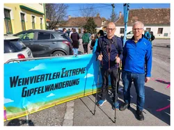 Zwei Männer stehen mit Trekkingstöcken auf einer Straße. Sie tragen blaue Jacken und Turnschuhe. Hinter ihnen befindet sich ein Banner mit schwarzem Text. Im Hintergrund gehen Menschen auf der Straße.