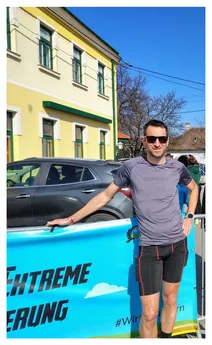 Ein Mann in einem Trainingsshirt und Sonnenbrille steht lächelnd neben einem blauen Banner. Er lehnt sich mit der linken Hand an eine Barriere. Hinter ihm steht ein geparktes Auto und ein Gebäude mit gelben Wänden und grünen Fenstern.