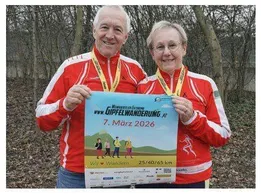 Zwei ältere Personen in passenden roten Jacken halten ein Banner für den Gippelwanderunning-Event am 7. März 2026. Sie tragen gelbe Medaillen und lächeln in einer Waldumgebung.