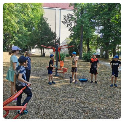 Eine Gruppe von Kindern und Erwachsenen versammelt sich um eine Spielplatzausrüstung in einem Park. Einige stehen, andere sitzen auf der Ausrüstung. Bäume und ein Gebäude sind im Hintergrund.