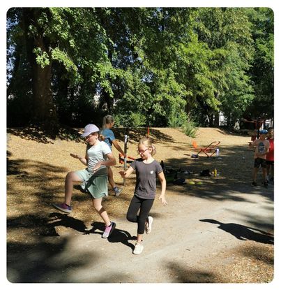 Zwei junge Mädchen rennen auf einem Pfad in einem Park mit Bäumen und Spielplatzgeräten im Hintergrund.