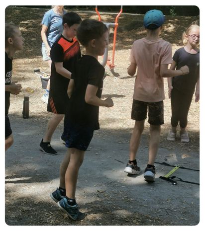 Fünf Kinder stehen in einem Park, zwei Jungen in schwarzen Shirts, einer in einer blauen Mütze, und zwei Mädchen, eines mit Brille. Sie scheinen an einer Aktivität beteiligt zu sein.