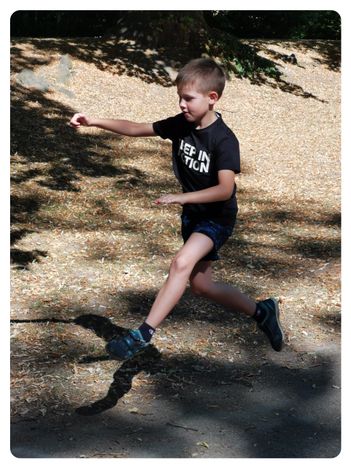 Ein junger Junge läuft in einem Park und trägt ein schwarzes T-Shirt mit den Worten 'Keep in Action' und blaue Shorts. Er ist barfuß und scheint in Bewegung zu sein.