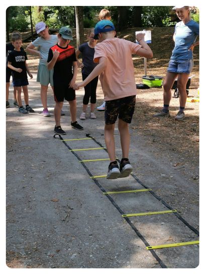 Eine Gruppe von Kindern übt auf einem Pfad Geschicklichkeitsübungen. Ein Kind springt über eine gelbe Leiter, während andere zuschauen.