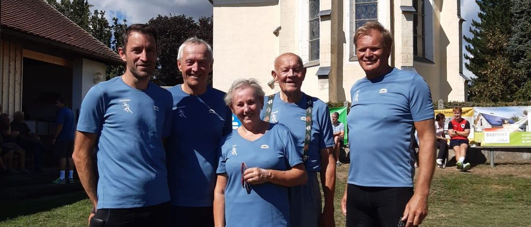 Fünf Personen, alle in passenden blauen T-Shirts und Shorts gekleidet, stehen gemeinsam vor einer Kirche mit einem roten Dach. Die Umgebung ist im Freien, mit einem Rasenbereich und einem klaren blauen Himmel.