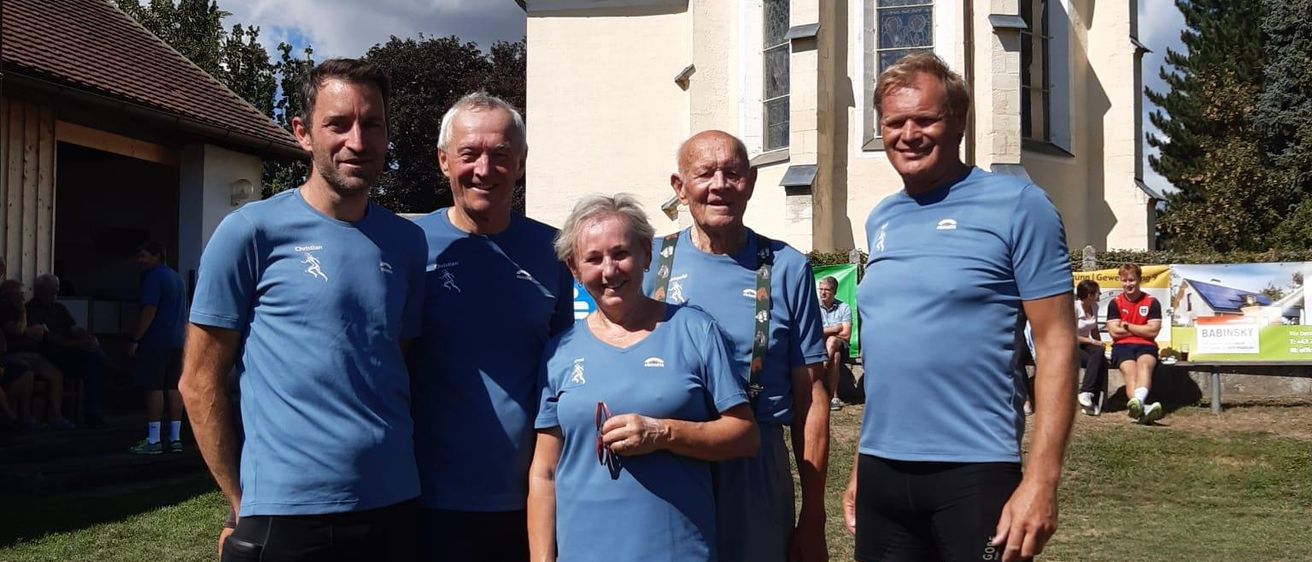 Fünf Personen, alle in passenden blauen T-Shirts und Shorts gekleidet, stehen gemeinsam vor einer Kirche mit einem roten Dach. Die Umgebung ist im Freien, mit einem Rasenbereich und einem klaren blauen Himmel.