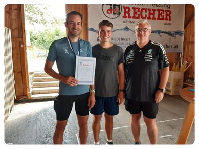 Drei Männer stehen nebeneinander, einer hält ein Zertifikat. Der Mann auf der linken Seite trägt ein blaues Shirt mit einer Medaille um den Hals. Der Mann in der Mitte trägt ein graues Shirt und hält ein Zertifikat. Der Mann auf der rechten Seite trägt ein schwarzes Shirt mit einem Logo auf der Brust. Hinter ihnen ist ein Banner mit dem Wort 'Recher'.