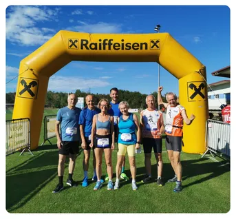 Eine Gruppe von sechs Läufern in Sportkleidung posiert für ein Foto unter einem gelben Raiffeisen-Bogen auf einem Rasenfeld.