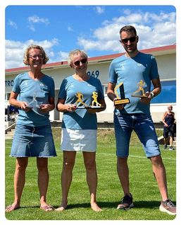 Drei Personen stehen auf einem Rasenfeld und halten Trophäen in der Hand. Zwei Frauen tragen blaue T-Shirts und Jeansröcke, während der Mann ein blaues T-Shirt und Jeansshorts trägt. Sie lächeln und posieren für ein Foto.