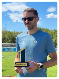 Ein Mann in einem blauen T-Shirt hält eine Trophäe mit der Aufschrift 'Steinbergkönig 2026' an einem sonnigen Tag auf einem Rasenfeld mit einem Fußballtor im Hintergrund.