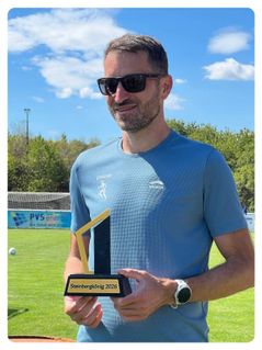 Ein Mann in einem blauen T-Shirt hält eine Trophäe mit der Aufschrift 'Steinbergkönig 2026' an einem sonnigen Tag auf einem Rasenfeld mit einem Fußballtor im Hintergrund.