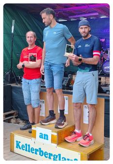 Drei Männer stehen auf einem Podium, zwei halten Trophäen. Der Mann in der Mitte trägt ein hellblaues Shirt und Shorts. Der Mann auf der linken Seite trägt ein rotes Shirt und Jeansshorts. Der Mann auf der rechten Seite trägt ein dunkelblaues Shirt und Shorts.