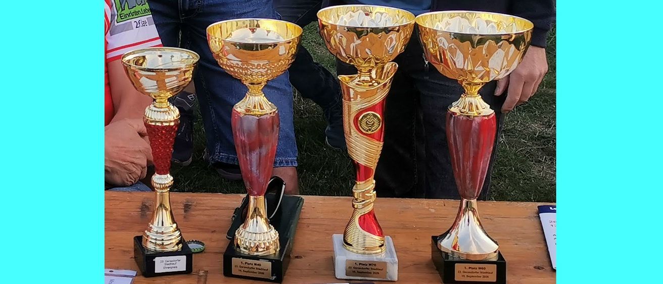 Vier goldene und rote Pokale sind auf einem Holztisch ausgestellt. Jeder Pokal hat ein einzigartiges Design und ein Etikett mit Text und Zahlen. Ein Mensch in Jeans steht hinter dem Tisch.