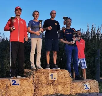 Fünf Personen stehen auf einem Strohballenpodium und halten Trophäen. Der Mann links trägt einen roten Mantel, die Frau neben ihm ein blaues Hemd. Der Mann in der Mitte trägt ein schwarzes Hemd.