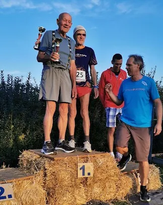 Eine Gruppe von Männern steht auf einem Strohballenpodium, einer von ihnen hält eine Trophäe und lächelt. Ein anderer Mann steht hinter ihm, und zwei weitere stehen zur Seite, einer trägt eine rote Jacke.