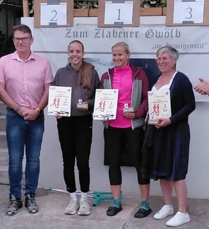 Vier Personen stehen lächelnd zusammen, einer Mann links und drei Frauen mit Urkunden. Sie stehen vor einem Banner mit den Nummern 1, 2 und 3.