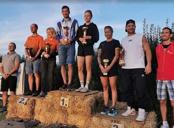 Eine Gruppe von Menschen steht auf Heuballen und hält Trophäen. Sie sind in Freizeitkleidung gekleidet und scheinen einen Sieg zu feiern. Der Ort ist im Freien mit einem klaren blauen Himmel im Hintergrund.