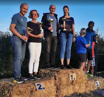 Eine Gruppe von Menschen steht auf einem Strohballenpodium, lächelt und hält Pokale. Sie sind lässig gekleidet und der Himmel ist klar.