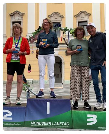 Vier Personen stehen auf einem Podest mit Medaillen in einer europäischen Stadt und lächeln für ein Foto. Jede trägt Laufausrüstung und hält eine Medaille. Das Podest ist von 1 bis 3 nummeriert.