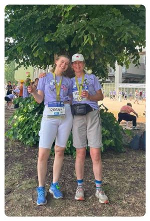 Zwei Frauen mit Medaillen um den Hals und Nummern auf ihren Trikots stehen unter einem Baum. Eine Person sitzt hinter ihnen auf dem Boden, und Menschen laufen im Hintergrund.