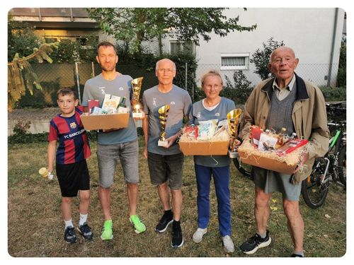 Eine Familie und Freunde versammeln sich draußen für ein Foto. Sie halten Geschenkkörbe und Trophäen. Der Mann in der Mitte trägt eine Brille. Bäume und ein Gebäude sind im Hintergrund.