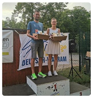 Ein Mann und eine Frau stehen auf einem Podest mit Geschenkboxen. Sie lächeln für ein Foto. Dahinter befindet sich ein Banner mit Text.