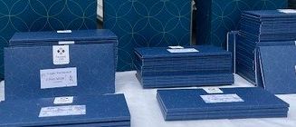 Ein Tisch zeigt mehrere blaue Geschenkboxen und Taschen mit weißen Etiketten und einem blauen Ballon in der Mitte. Jede Tasche und jedes Kästchen trägt den Schriftzug 'THERME LAA'. Der Hintergrund zeigt eine Treppe mit Metallgeländern.
