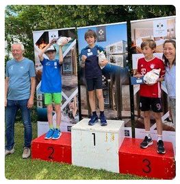 Drei Jungen stehen mit Medaillen auf einem Podest, zwei von ihnen halten Trophäen. Einer trägt ein blaues Shirt und grüne Shorts, der andere ein blaues Shirt und schwarze Shorts. Der dritte Junge trägt ein rotes Trikot und schwarze Shorts. Ein Mann in einem blauen Shirt steht hinter ihnen.