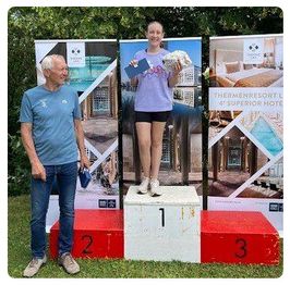 Ein älterer Mann und eine junge Frau stehen auf einem Podium mit der Nummer 1. Die Frau hält eine Trophäe und einen Blumenstrauß. Dahinter stehen Banner mit Logos.
