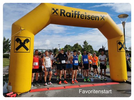 Läufer stellen sich am Start eines Rennens auf, tragen nummerierte Startnummern und stehen unter einem gelben Bogen, auf dem Raiffeisen steht.