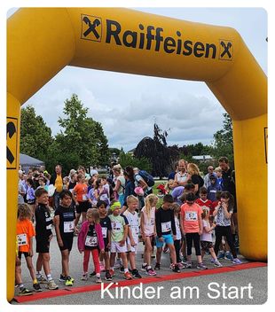 Kinder stehen in einer Reihe unter einem gelben Bogen mit der Aufschrift Raiffeisen, bereit für einen Lauf. Einige tragen Nummern auf ihren Trikots.