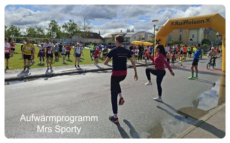 Läufer nehmen an einem Marathon teil, mit einem Bogen, auf dem Raiff steht und die Marke Mrs. Sporty sichtbar ist. Teilnehmer laufen auf einer Straße, während Zuschauer von der Seite zuschauen.