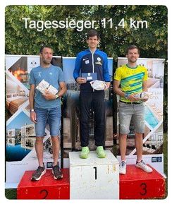 Drei Männer stehen auf einem Podest, halten Trophäen und Taschen. Der Mann in der Mitte trägt ein blaues und gelbes Trikot, während die anderen lässige Kleidung tragen. Dahinter ist eine Kulisse mit einem Bild eines Gebäudes und Bäumen.