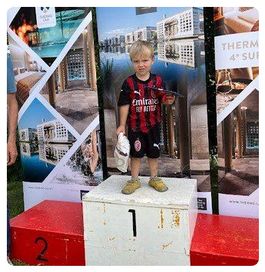 Ein junger Junge in einem Fußballtrikot steht auf einem Podium mit einem Pokal und einer Plastiktüte und hält einen Bogen und einen Pfeil. Hinter ihm sind Poster mit Gebäuden und Landschaften.
