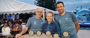 Drei Personen stehen hinter einem Tisch mit Medaillen. Der Mann links trägt eine Brille und ein blaues Shirt. Die Frau in der Mitte trägt eine Brille und eine blaue Jacke. Der Mann rechts trägt ein blaues Shirt. Dahinter steht ein Zelt.