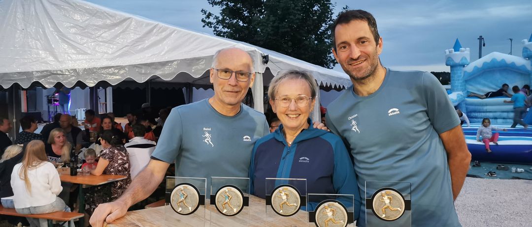 Drei Personen stehen hinter einem Tisch mit Medaillen. Der Mann links trägt eine Brille und ein blaues Shirt. Die Frau in der Mitte trägt eine Brille und eine blaue Jacke. Der Mann rechts trägt ein blaues Shirt. Dahinter steht ein Zelt.