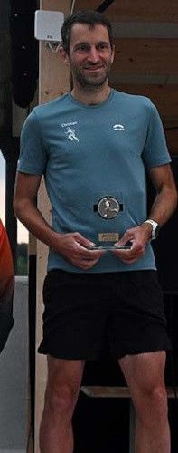 Ein Mann steht lächelnd und hält eine Trophäe in den Händen, er trägt ein blaues T-Shirt und schwarze Shorts.